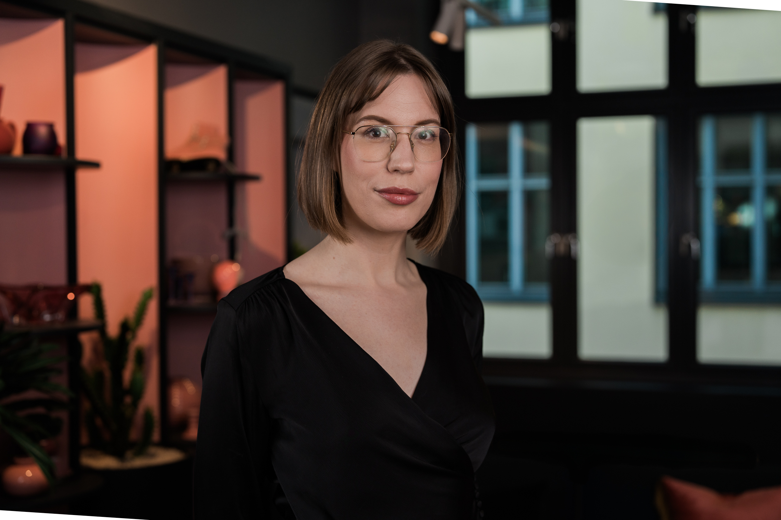 A person with short brown hair and glasses is standing indoors in front of a shelf with decorative items in warm colors. The person is wearing a black top and is looking directly at the camera with a neutral expression. The background is subdued and features large windows and modern interior design