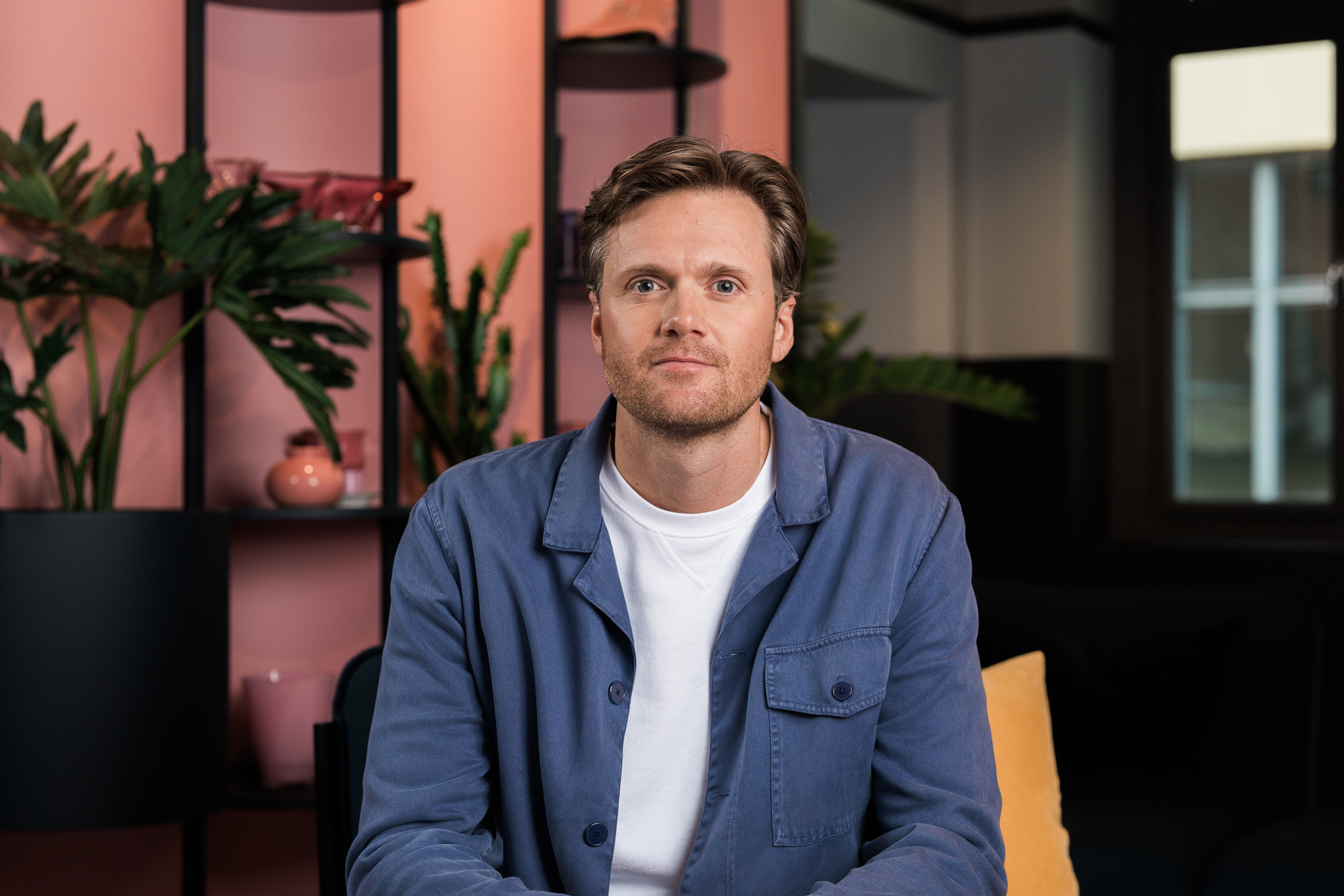 Eirik Dysthe, strategisjef i FINN eiendom, sitter i et moderne kontormiljø med rosa bakvegg, grønne planter og myk belysning, og ser rolig mot kameraet.