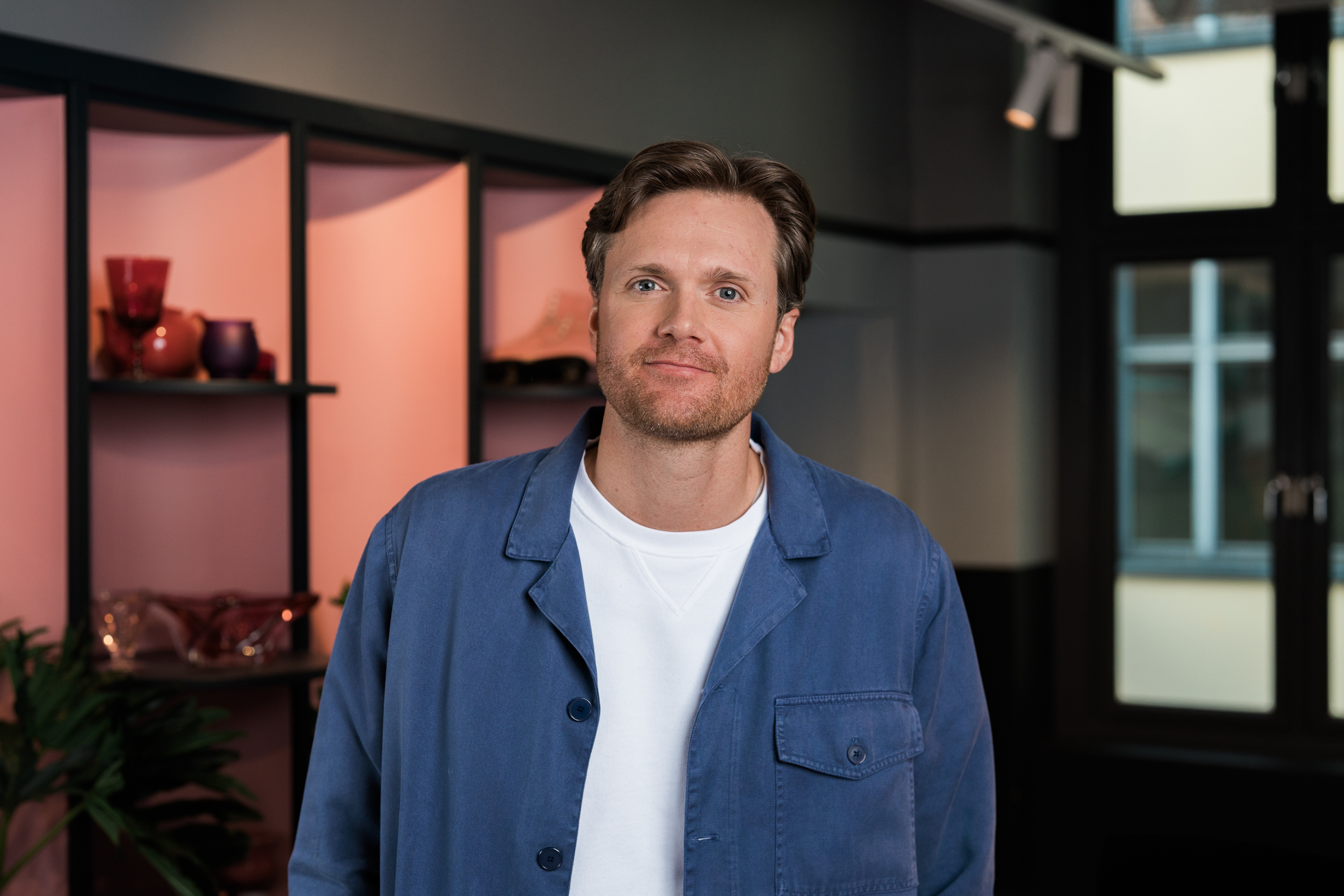 En mann med kort, brunt hår og skjegg står innendørs foran hyller med dekorative gjenstander. Han har på seg en blå jakke over en hvit genser og ser rolig inn i kameraet. Bakgrunnen er et moderne rom med pastellfargede hyller og store vinduer.