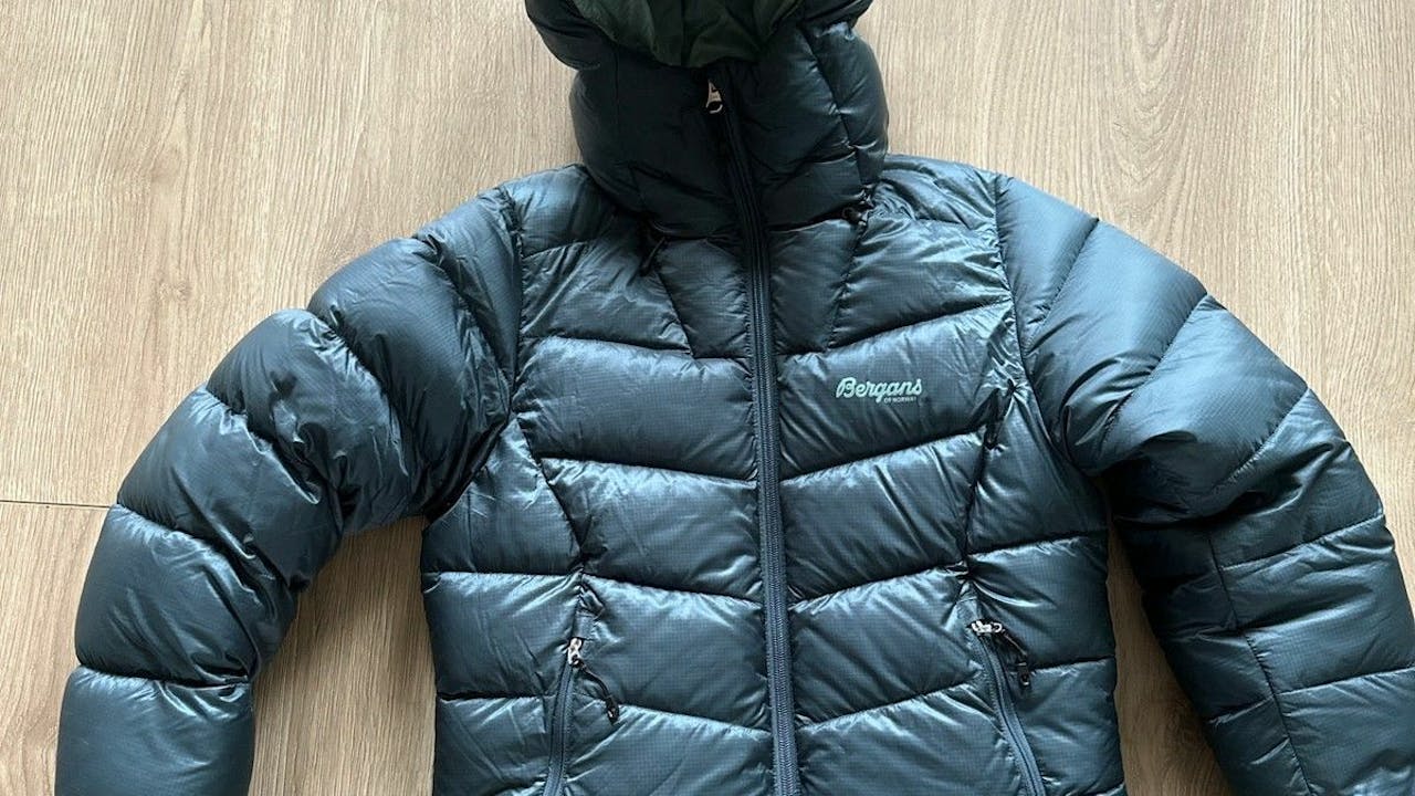 Mørk grønn Bergans dunjakke med hette og glidelås, liggende flatt på et lyst tregulv.
