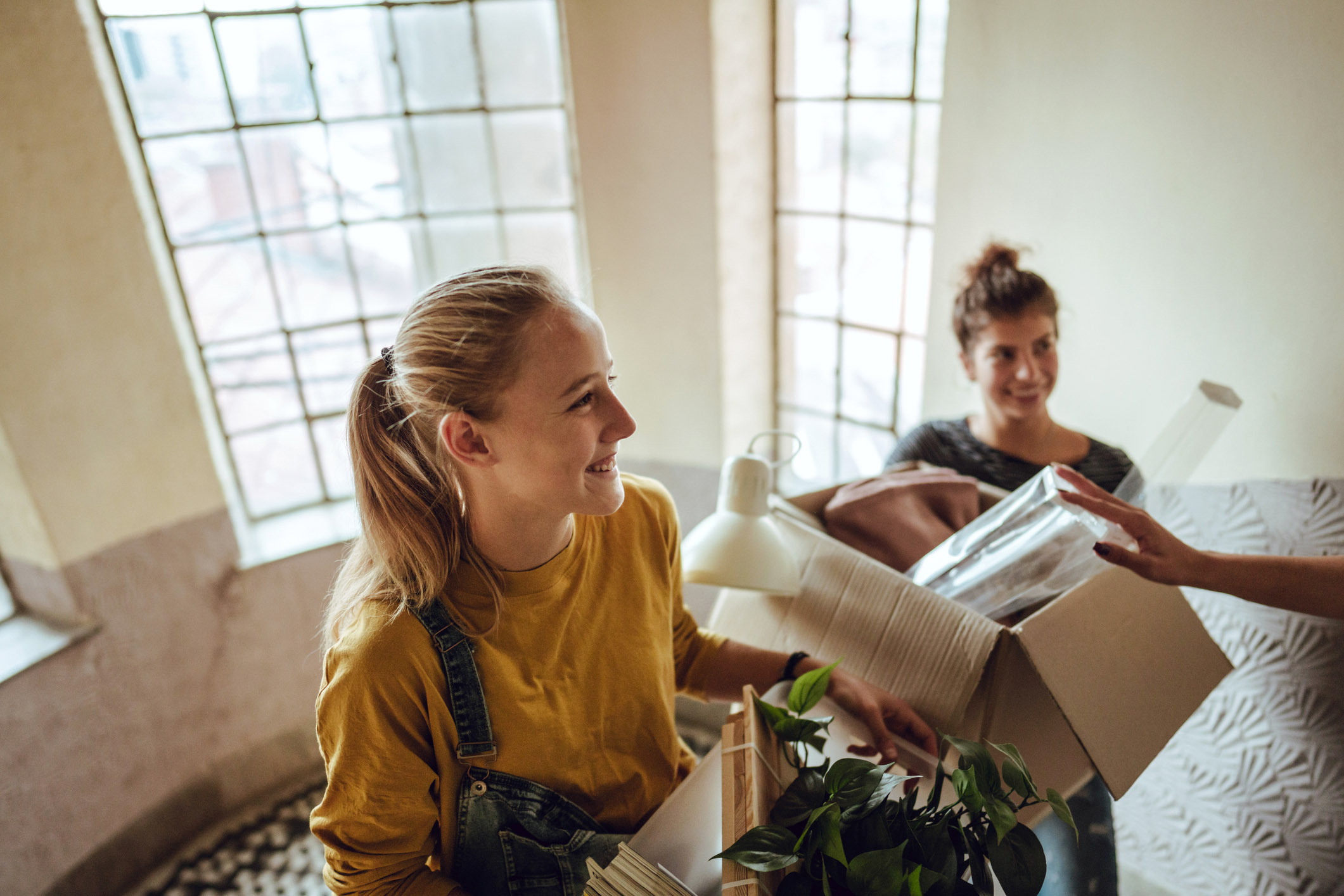 To unge kvinner flytter inn i en ny bolig, smilende mens de bærer esker og husholdningsartikler opp en trapp med store vinduer i bakgrunnen. Atmosfæren er lett og positiv, og gjenspeiler gleden over en ny start.