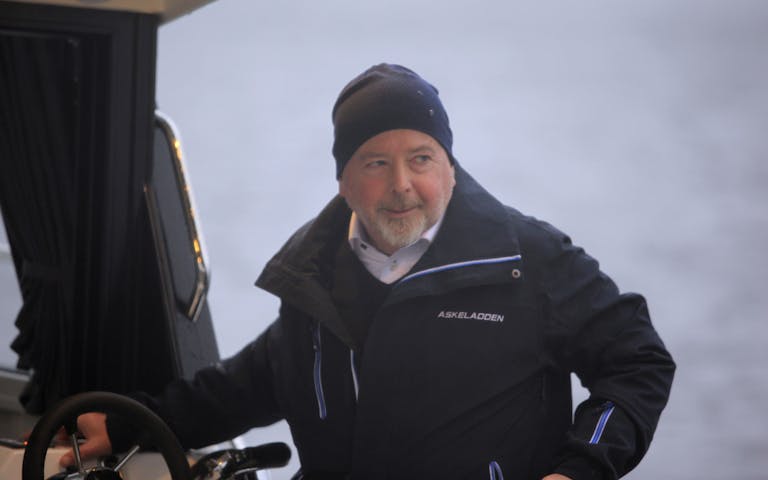 Bildet viser Henrik Askvik, daglig leder hos Askeladden Boats, ikledd sort regnjakke og lue, som kjører båt.