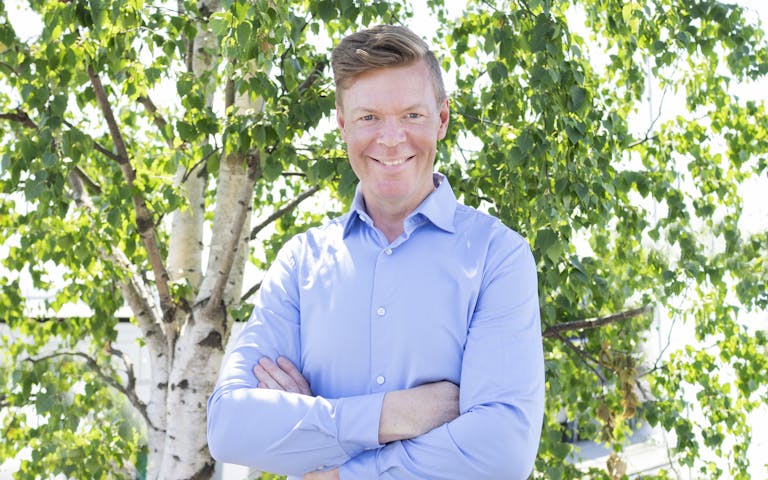 Bransjeansvarlig for FINN næring, Per Arne Berg-Heggelund, poserer smilende i blå skjorte foran grønt løvverk.