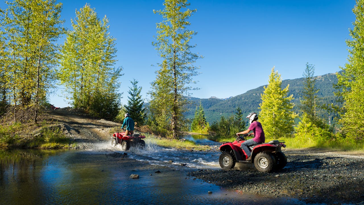 På bildet ser man blå himmel og sol, grønne trær og to personer på hver sin røde ATV (firhjuling), som kjører over en bekk.