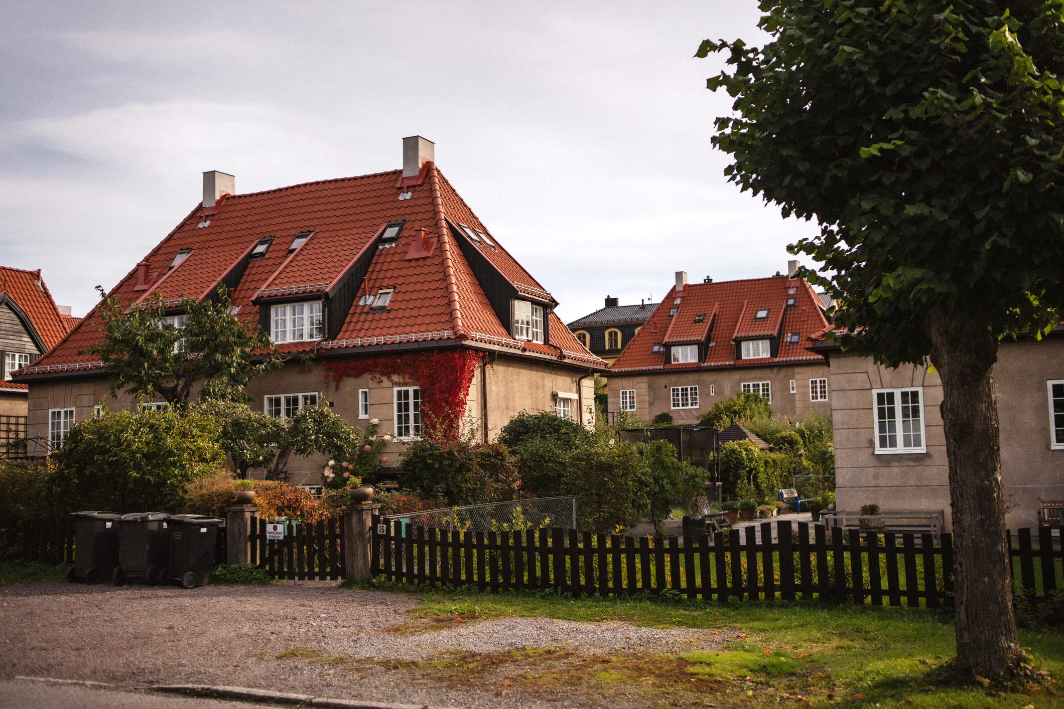 Et boligområde i Ullevål hageby med murhus og røde takstein omgitt av hager, trær og et mørkt tregjerde i forgrunnen.