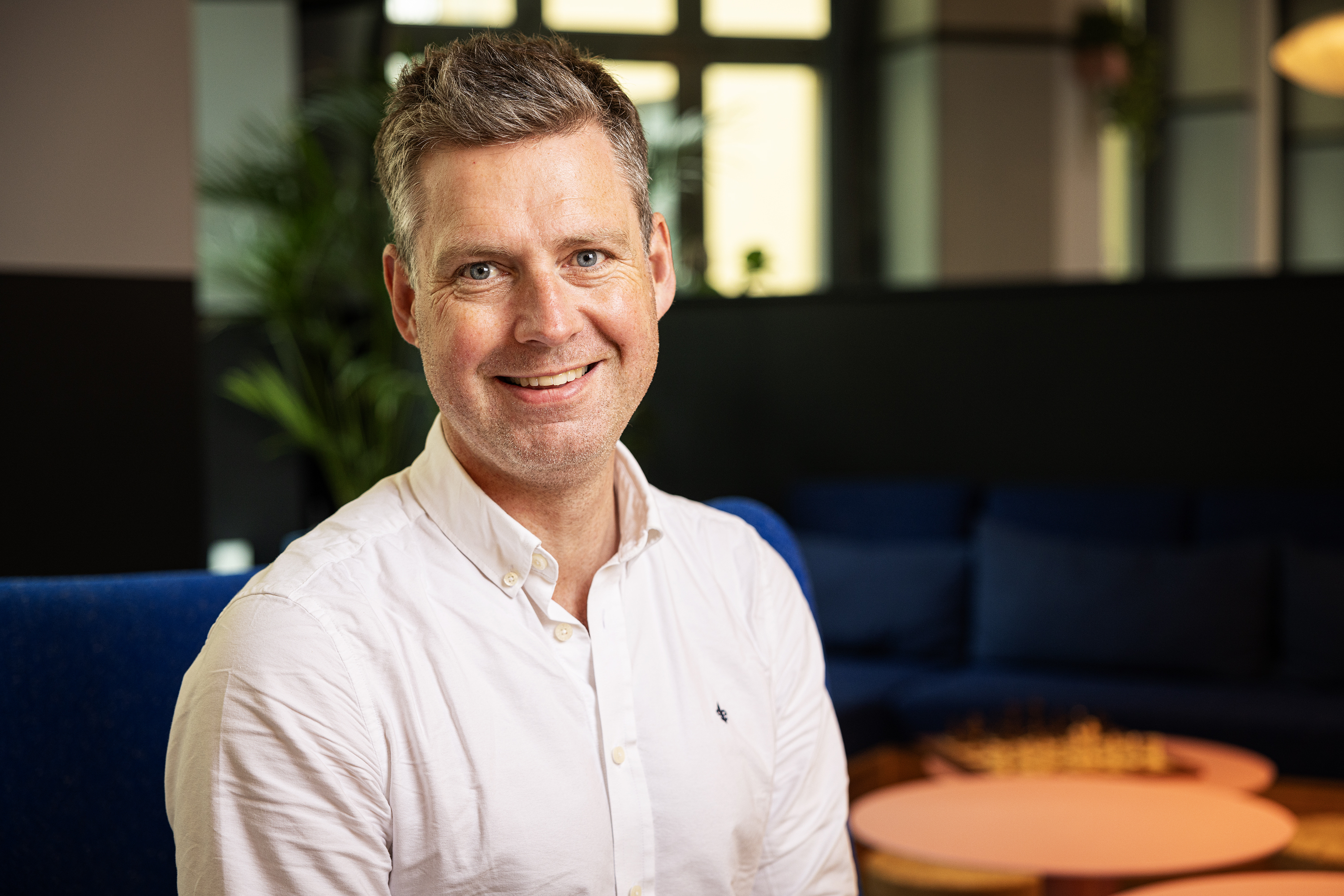En mann med kort, mørkt hår og iført en hvit skjorte smiler til kameraet mens han sitter i en komfortabel stol i et moderne innendørsmiljø.