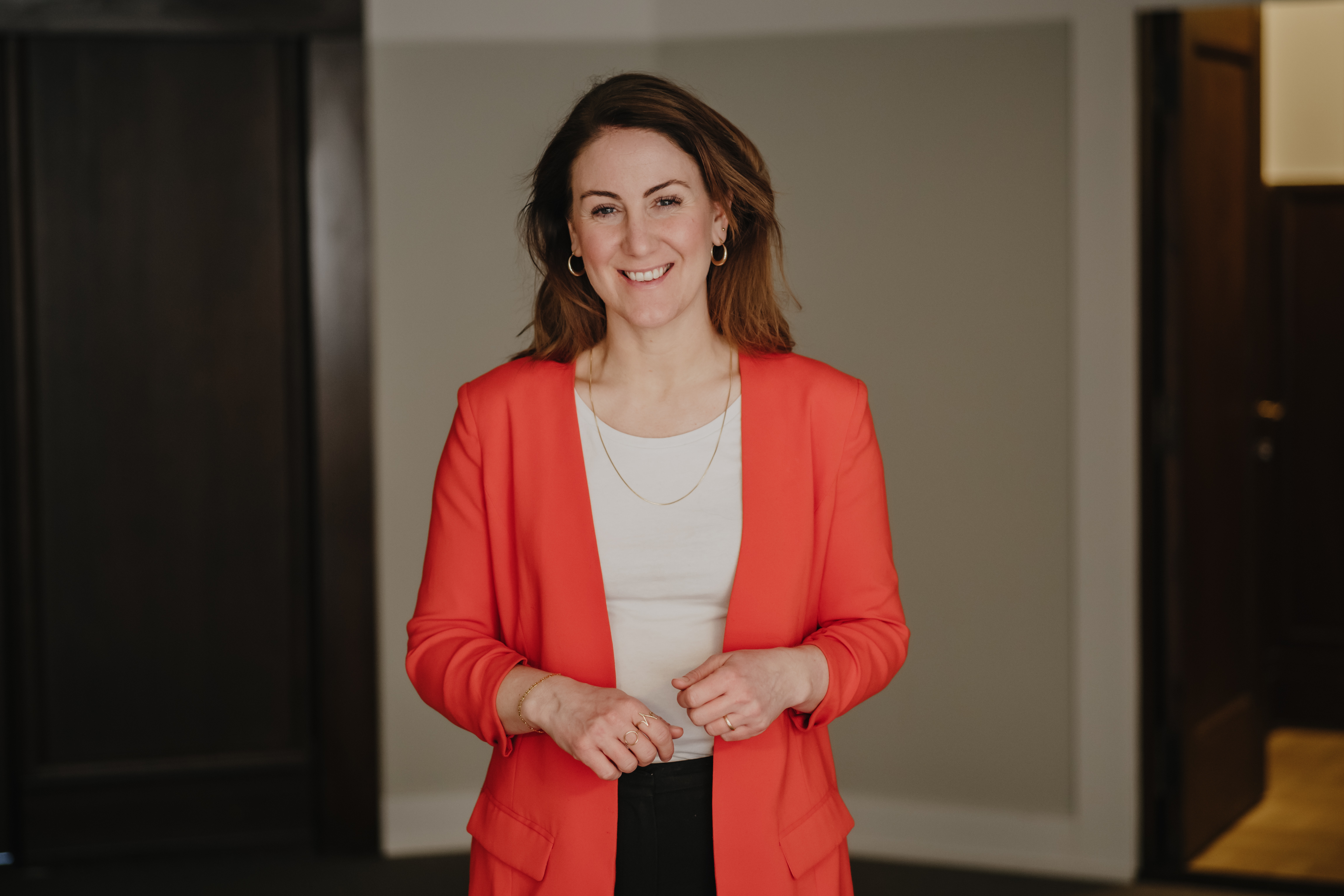 Arbeids- og inkluderingsminister Tonje Brenna med skulderlangt, brunt hår står smilende iført en rød blazer og hvit topp, med hendene samlet foran seg, i et rom med mørke dører og lyse vegger i bakgrunnen
