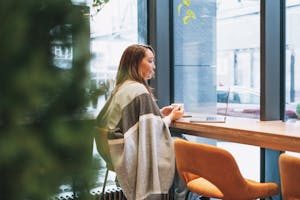 Kvinne sitter på cafe med kaffe og laptop. Ser tenkende ut.