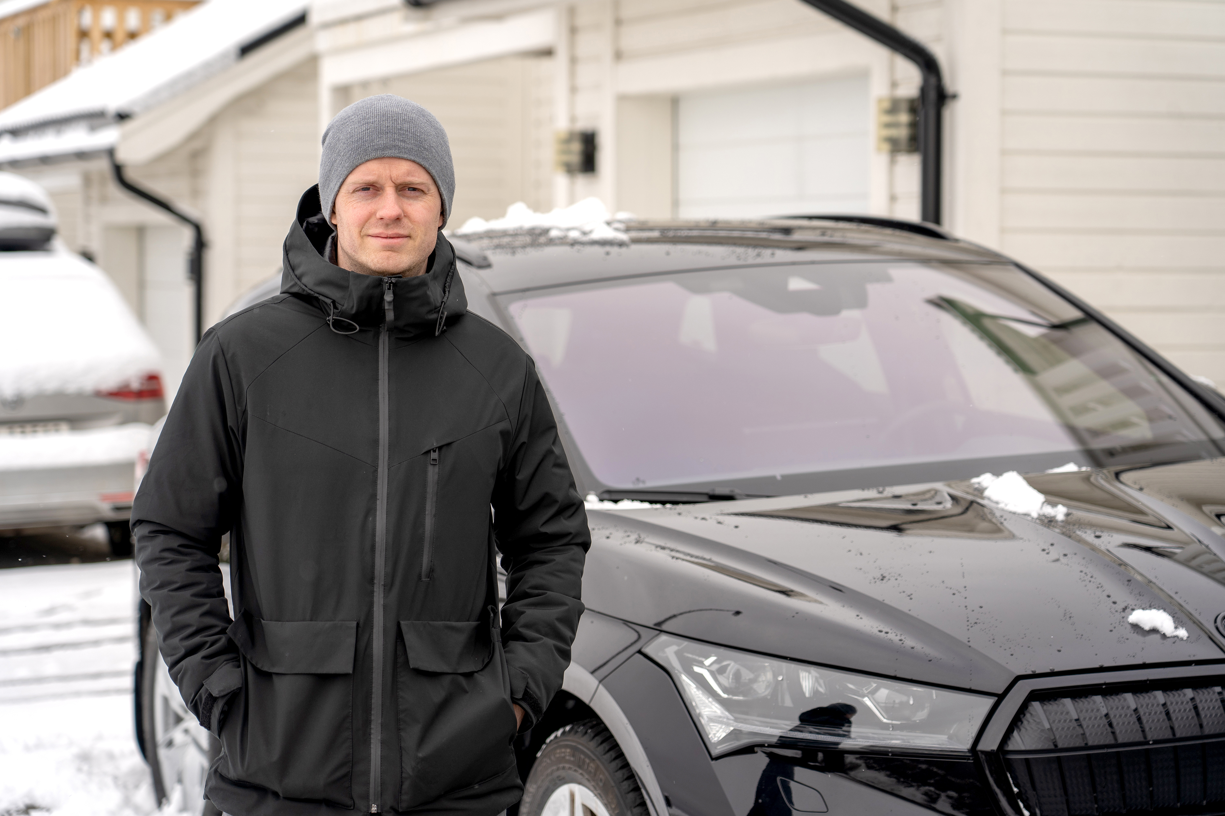 Mann med sort jakke og grå lue står foran sort elbil og ser i kamera