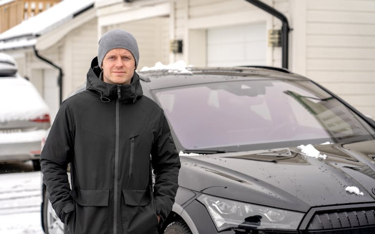 Mann med sort jakke og grå lue står foran sort elbil og ser i kamera