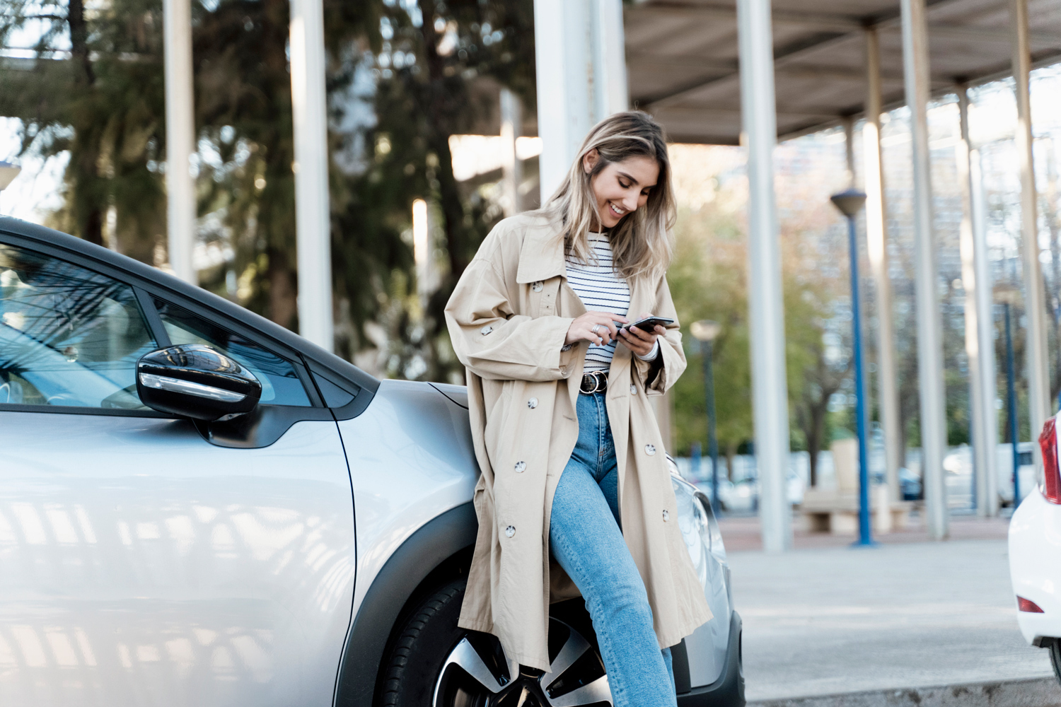 Dame står smilende ved bilen sin, mens hun ser på telefonen.