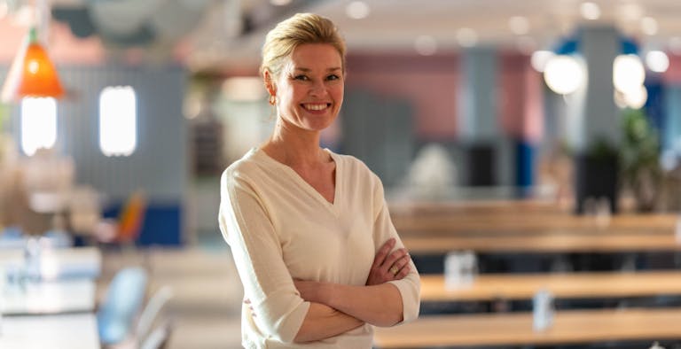 A confident middle-aged Caucasian woman with short blonde hair and a bright smile stands with her arms crossed. She is wearing a white V-neck blouse and is positioned in a spacious office environment with colorful decor and furniture in the background, suggesting a modern and casual workplace.
