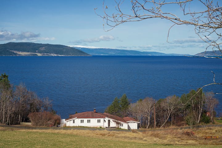 A scenic view of a large, serene blue lake with a white house in the foreground. The house is surrounded by a grassy field and scattered trees. In the distance, rolling hills and snow-capped mountains line the far side of the lake under a clear blue sky.