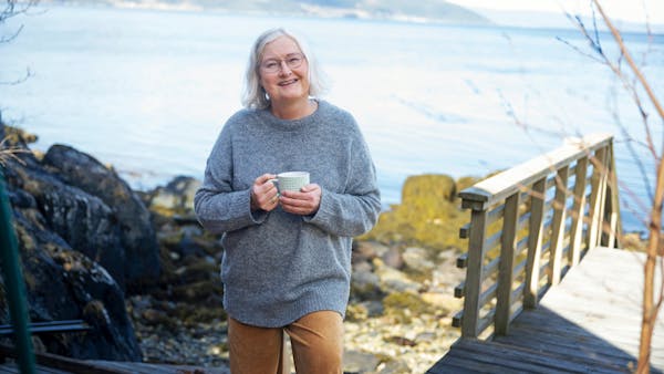 An elderly woman with glasses and gray hair smiles warmly while holding a mug. She is standing on a wooden walkway by a lakeside, wearing a cozy gray sweater and tan pants. In the background, a calm lake and distant mountains create a serene atmosphere.